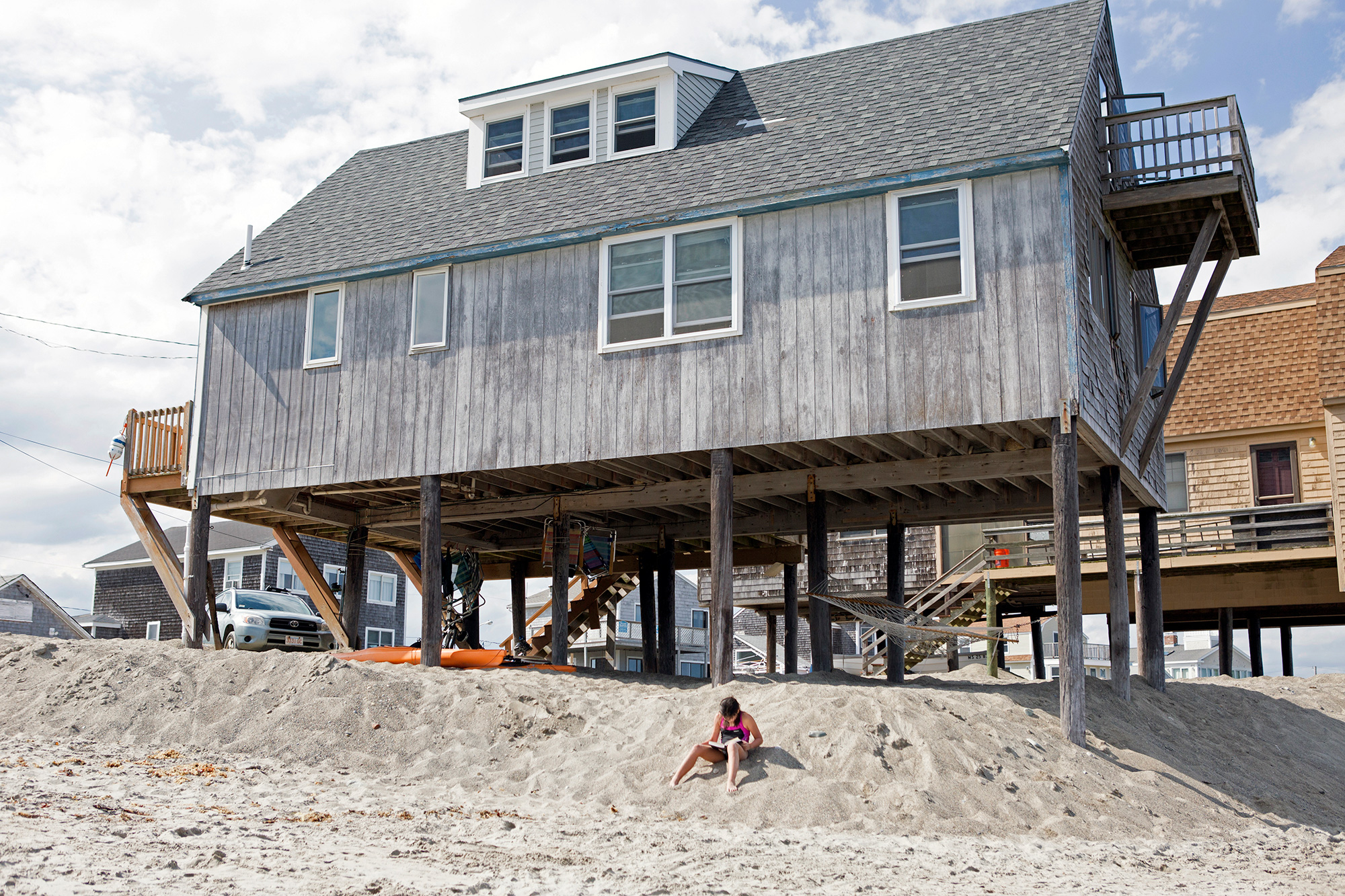 A girl reads her book by a raised cottage on Peggotty Beach in Scituate, on the South Shore of Massachusetts. Where there were once more than 50 cottages, the number has dwindled to a dozen as storms have eroded the beach and forced homeowners to vacate the area. Those houses that remain have been moved onto stilts. (Photo by Lauren Owens Lambert/GroundTruth)