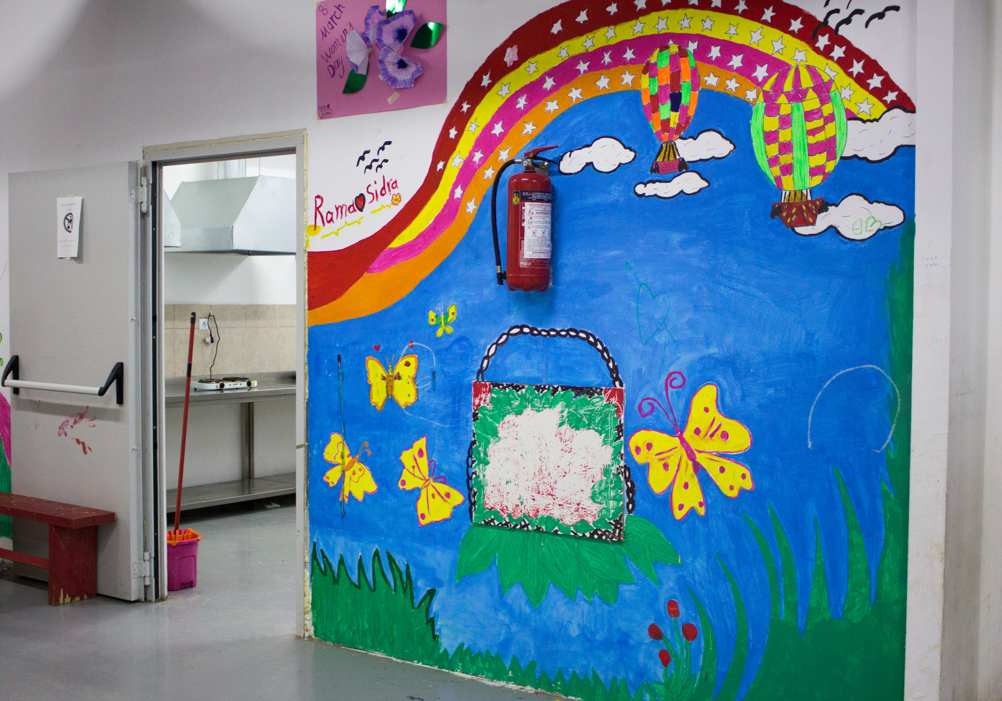 Throughout Elpida Home, murals line the walls. The communal kitchen is around the corner of this fluorescent mural. Residents can make their own meals and chat through the window that peers out into the tea room. (Photo by Catherine Clark/GroundTruth)