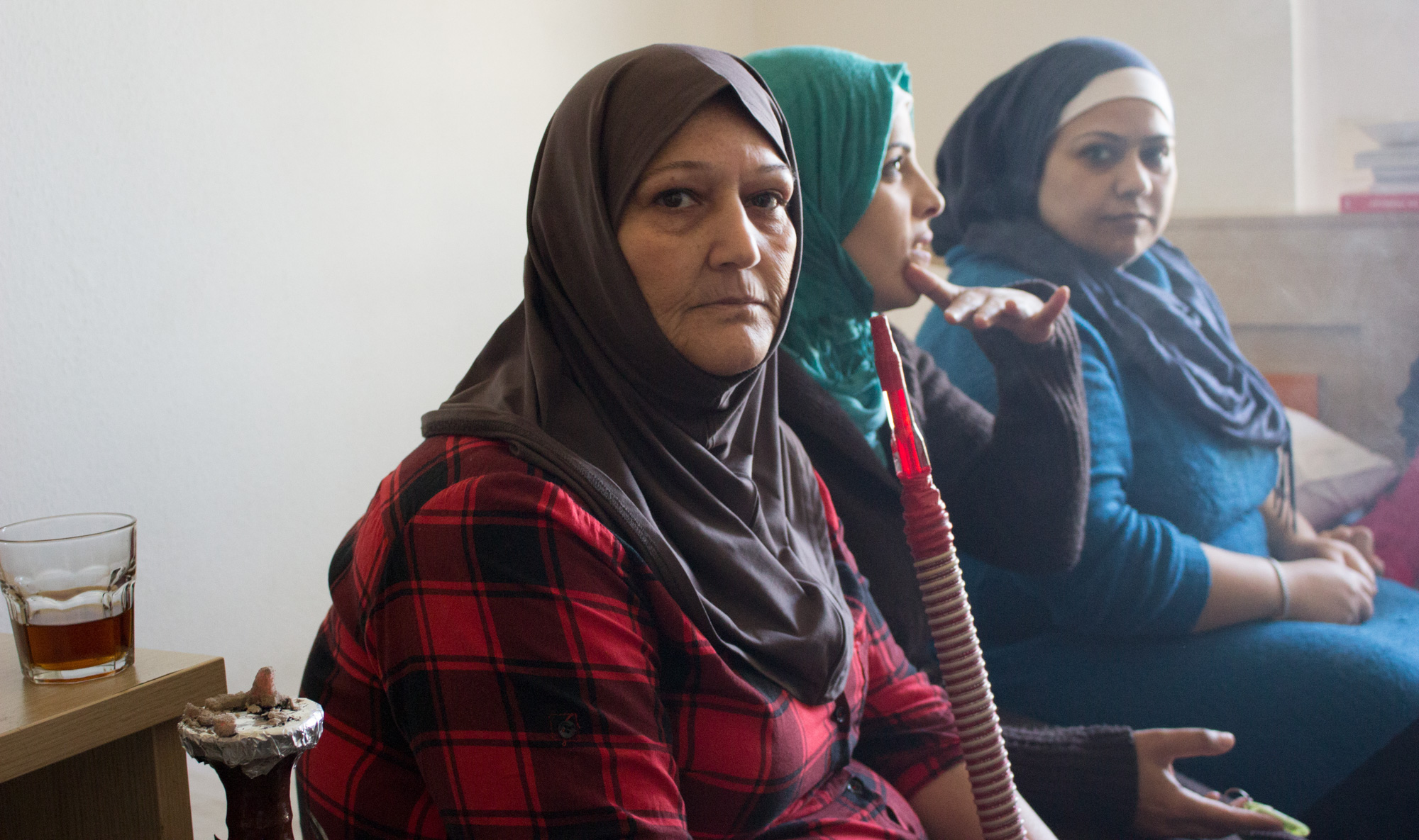 Grasping a hookah pipe, Syrian refugee Hanifa Hussein sits on a twin bed with her older daughter (right) and family friend (left). Smoking is a common way adult residents pass time at the Sindos apartment complex outside Thessaloniki, Greece (Photo by Catherine Clark/GroundTruth) 