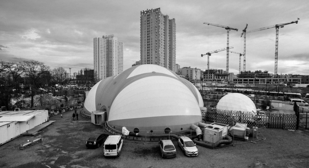 The Dome theatre is visible to the right of La Bulle, the shelter for migrants run by Emmas Solidarat at Porte de La Chapelle in Paris's 18th arrondissement. Both structures are located in an old rail yard in the north of the city. Most of the migrants and refugees who are granted shelter here spent several nights sleeping on the streets in and around Port de la Chapelle before being allowed in, due to the limited space available in La Bulle's accommodation center. (Photo by Annabelle Marcovici/GroundTruth)